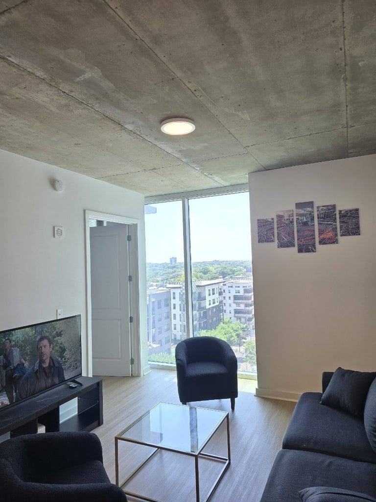 Modern high-rise apartment living room with concrete ceiling, black seating, TV, and floor-to-ceiling windows overlooking city skyline